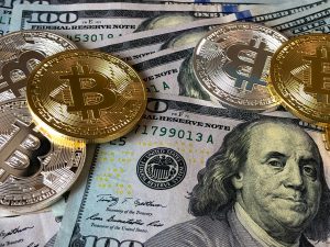 Golden Bitcoin commemorative coins scattered across a collection of US dollar bills including hundred-dollar bills featuring Benjamin Franklin, representing the intersection of traditional seed funding and cryptocurrency investment. The metallic Bitcoin coins with their distinctive 'B' logos and concentric circle patterns contrast against the green and blue Federal Reserve notes, symbolizing the evolving landscape of startup financing and digital assets in modern seed funding rounds.