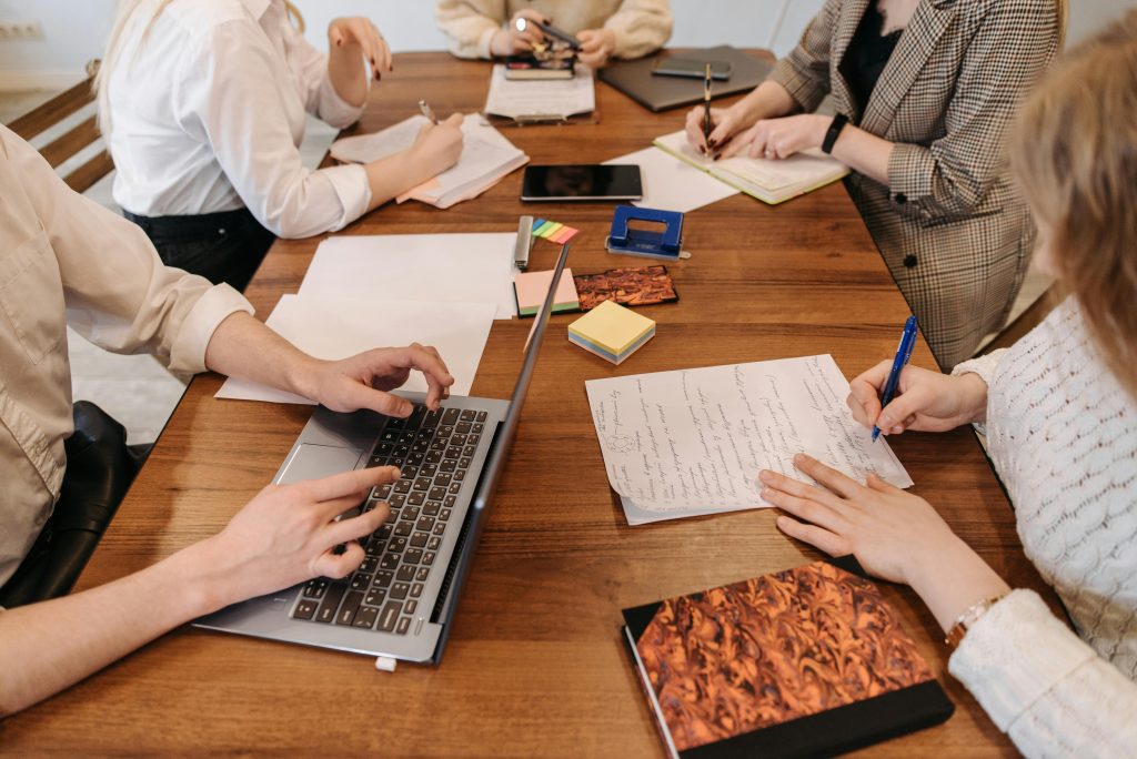 This image shows a startup idea validation session with team members collaborating around a wooden table. They're using a mix of digital tools (laptops, tablets) and traditional methods (handwritten notes, documents) to work through their concepts. The scene captures the typical brainstorming environment where entrepreneurs gather to test and refine their business ideas using both analog and digital approaches.