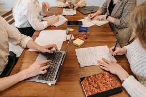 This image shows a startup idea validation session with team members collaborating around a wooden table. They're using a mix of digital tools (laptops, tablets) and traditional methods (handwritten notes, documents) to work through their concepts. The scene captures the typical brainstorming environment where entrepreneurs gather to test and refine their business ideas using both analog and digital approaches.