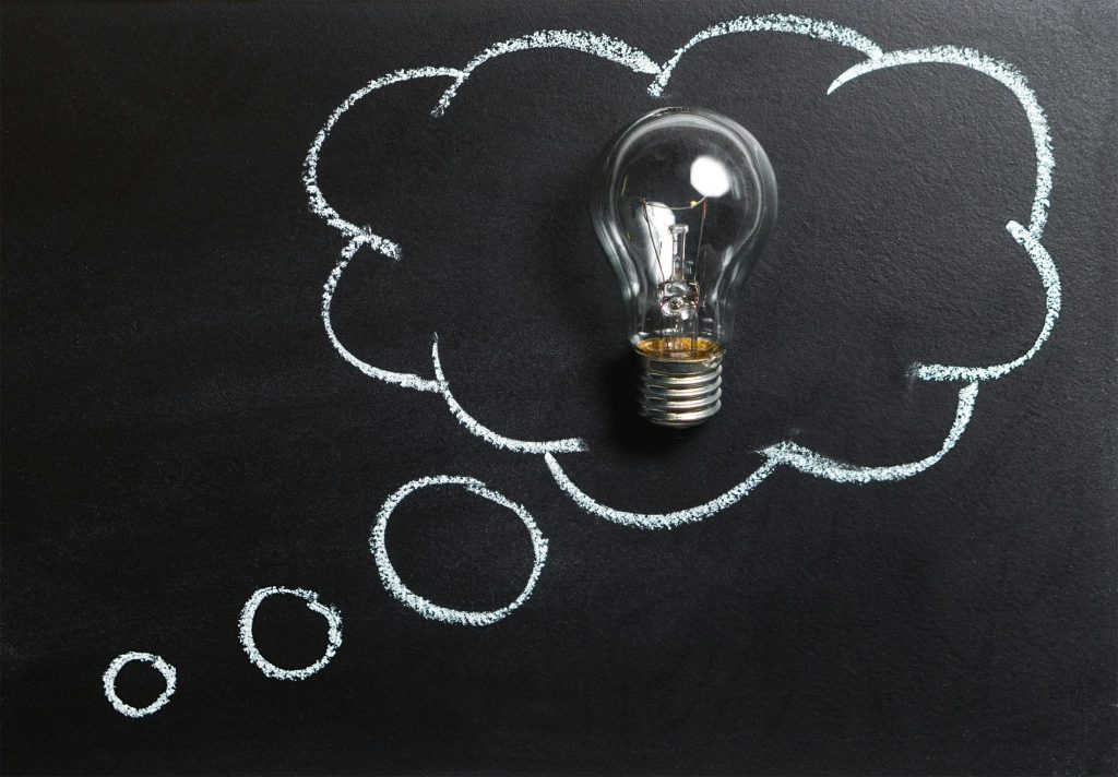 A glowing light bulb sits within a white chalk-drawn thought bubble on a dark blackboard surface, symbolizing the eureka moment of startup idea validation. The illuminated bulb represents the breakthrough when entrepreneurs validate their business concepts, transforming abstract thoughts into viable startup opportunities. Additional smaller circles trail from the thought bubble, illustrating the iterative process of refining and validating ideas before moving forward with seed funding and development.