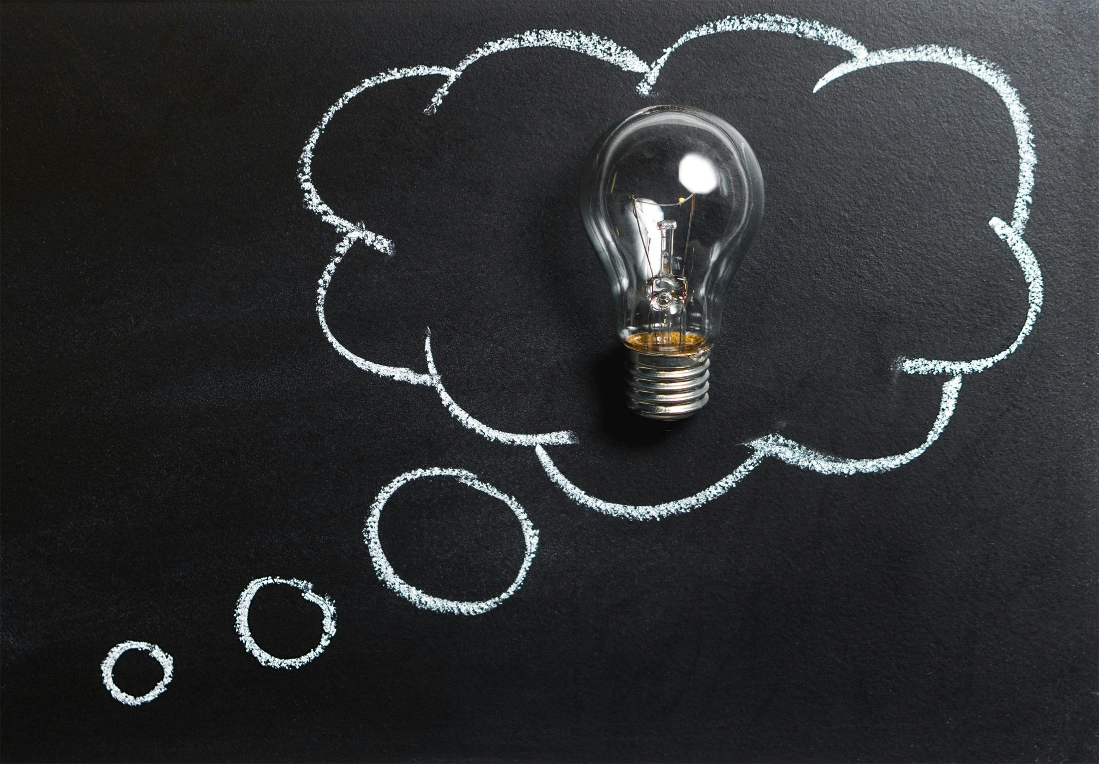 A glowing light bulb sits within a white chalk-drawn thought bubble on a dark blackboard surface, symbolizing the eureka moment of startup idea validation. The illuminated bulb represents the breakthrough when entrepreneurs validate their business concepts, transforming abstract thoughts into viable startup opportunities. Additional smaller circles trail from the thought bubble, illustrating the iterative process of refining and validating ideas before moving forward with seed funding and development.