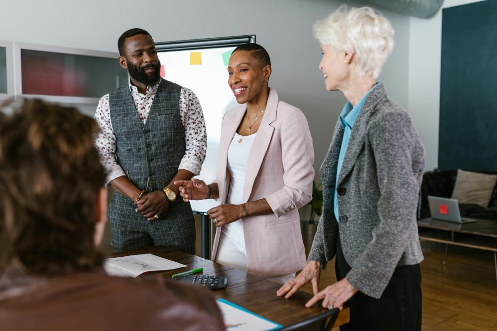 A professional business meeting scene showing four people in an office setting, likely including investors and entrepreneurs. In the foreground, a person with curly hair is partially visible from behind. Three colleagues are standing and engaged in conversation around a conference table: a Black man wearing a patterned shirt with a gray vest, a Black woman in a light pink blazer over a white top, and an older woman with short white/gray hair wearing a gray blazer with a blue accent. The woman in pink appears to be presenting or pitching while the others listen attentively, suggesting this could be an investor meeting or business pitch session. The modern office environment features a whiteboard with colorful sticky notes in the background, papers and documents on the wooden table, and contemporary office lighting.